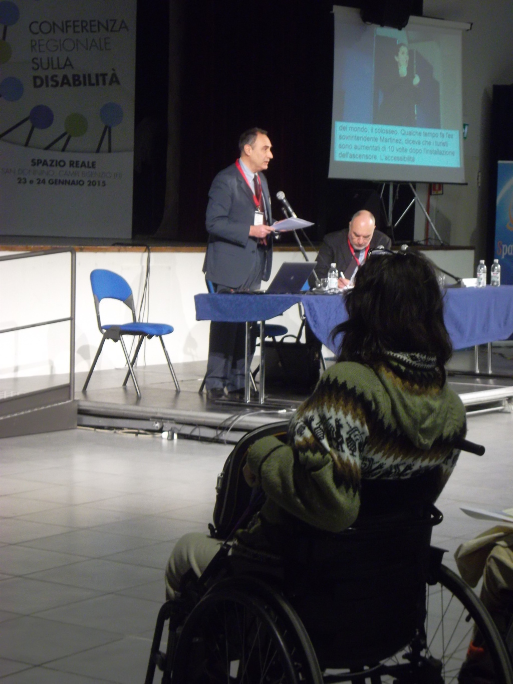 Tavolo relatori con Antonio Laurìa che parla e Emanuele Rossi, in primo piano vista da dietro donna su sedia a ruote