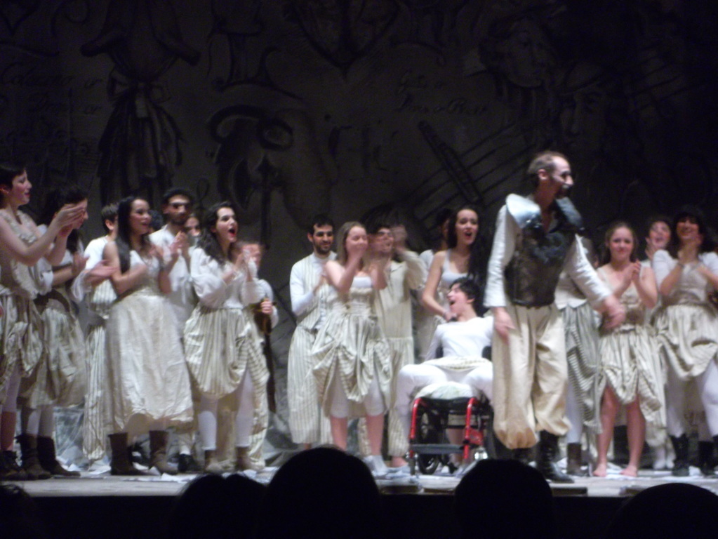 Foto momento dello spettacolo teatrale serale, Don Chisciotte con armatura e attori del coro in bianco che applaudono